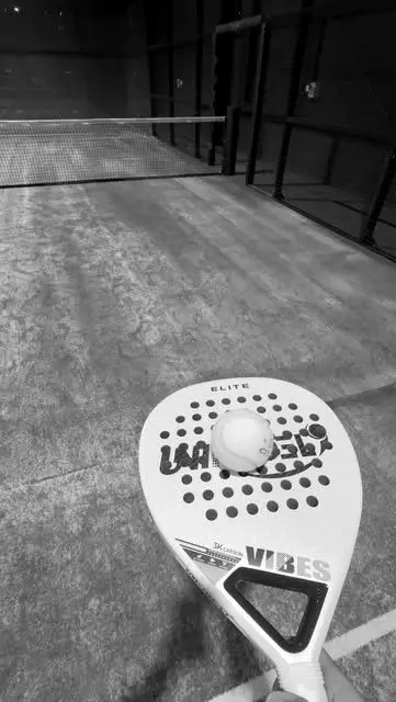 A padel bat resting on the court floor with a ball against the strings.